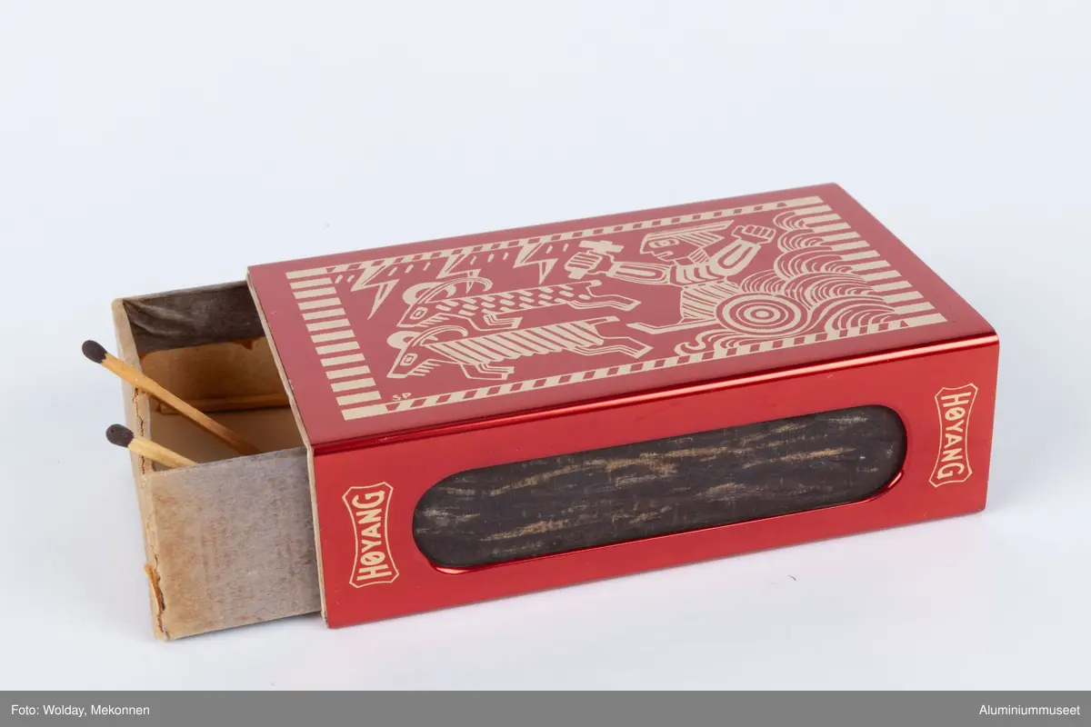 Rød, eloksert aluminium, med motiv Tor med hammeren.

Teknikk: En Aluminiumsplate ble hjørnestanset (avrundet) og to lange hull for ripeflate ble utstanset. Holderen ble knekket i to operasjoner i eksenterpresse. Først ble bunn knekket i en operasjon og så ble topp/side knekket i en operasjon. Begge operasjonene ble utført i eksenterpresse. Holderen ble polert, eloksert og farvet gul. Motivet " Tor med hammeren" ble siketrykket på med et dekkskjikt. Holderen ble så rødfarvet, men rødfarven angrep ikke motivet som var dekket. Eloksskiktet ble tilslutt tettet i sealeren (varmt vann). 
Form: Tilpasset utvendig form for stor fyrstikkeske