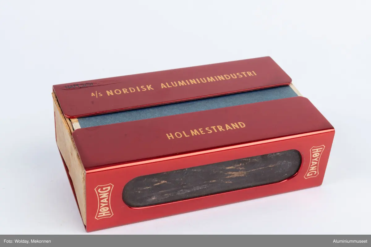 Rød, eloksert aluminium, med motiv Tor med hammeren.

Teknikk: En Aluminiumsplate ble hjørnestanset (avrundet) og to lange hull for ripeflate ble utstanset. Holderen ble knekket i to operasjoner i eksenterpresse. Først ble bunn knekket i en operasjon og så ble topp/side knekket i en operasjon. Begge operasjonene ble utført i eksenterpresse. Holderen ble polert, eloksert og farvet gul. Motivet " Tor med hammeren" ble siketrykket på med et dekkskjikt. Holderen ble så rødfarvet, men rødfarven angrep ikke motivet som var dekket. Eloksskiktet ble tilslutt tettet i sealeren (varmt vann). 
Form: Tilpasset utvendig form for stor fyrstikkeske