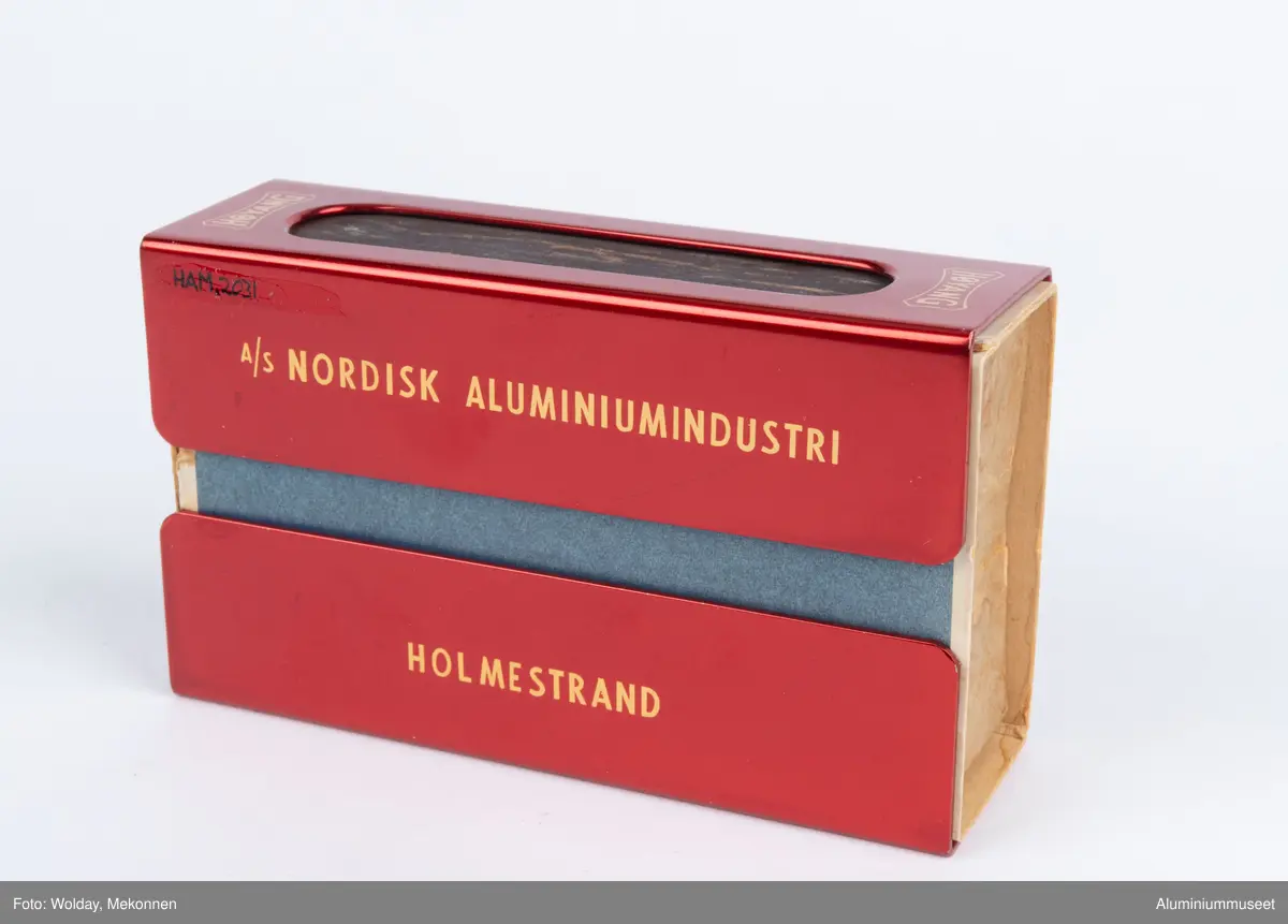 Rød, eloksert aluminium, med motiv Tor med hammeren.

Teknikk: En Aluminiumsplate ble hjørnestanset (avrundet) og to lange hull for ripeflate ble utstanset. Holderen ble knekket i to operasjoner i eksenterpresse. Først ble bunn knekket i en operasjon og så ble topp/side knekket i en operasjon. Begge operasjonene ble utført i eksenterpresse. Holderen ble polert, eloksert og farvet gul. Motivet " Tor med hammeren" ble siketrykket på med et dekkskjikt. Holderen ble så rødfarvet, men rødfarven angrep ikke motivet som var dekket. Eloksskiktet ble tilslutt tettet i sealeren (varmt vann). 
Form: Tilpasset utvendig form for stor fyrstikkeske