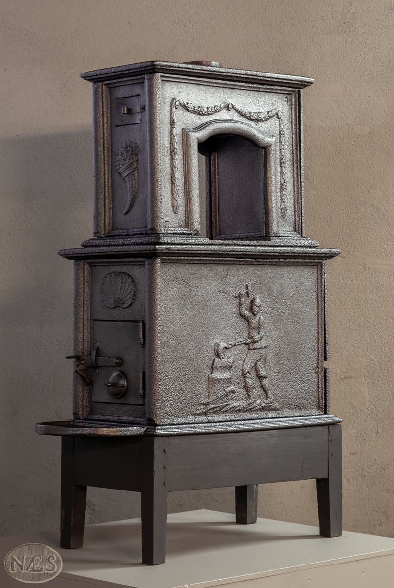 Etasjeovn med ovnsluke på kortsiden og røykuttak i toppen. På langsideplaten 1. etg en smed som holder en tang og hammer. En ambolt og et sverd liggende på bakken. Langside 2. etg har en buet åpning med girlande hengende rundt. Kortsiden 1. etg har en musling øverst. På kortsidene 2. etg et overflødighetshorn. Alle flatene er glate.