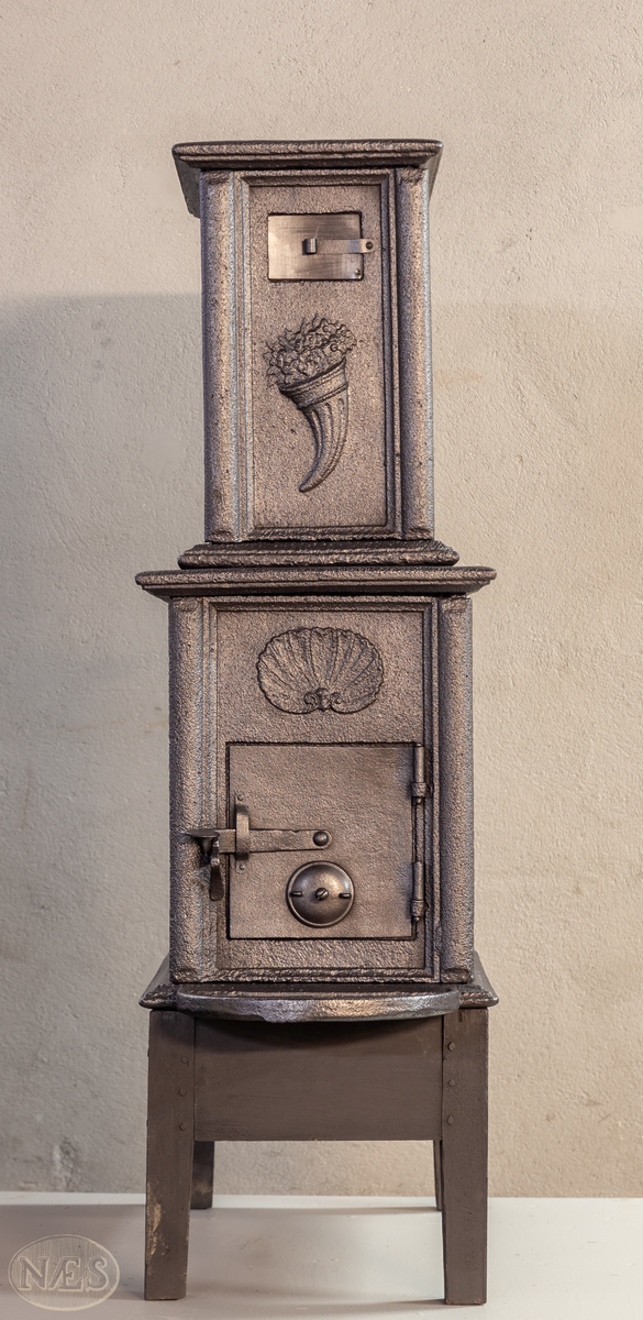 Etasjeovn med ovnsluke på kortsiden og røykuttak i toppen. På langsideplaten 1. etg en smed som holder en tang og hammer. En ambolt og et sverd liggende på bakken. Langside 2. etg har en buet åpning med girlande hengende rundt. Kortsiden 1. etg har en musling øverst. På kortsidene 2. etg et overflødighetshorn. Alle flatene er glate.