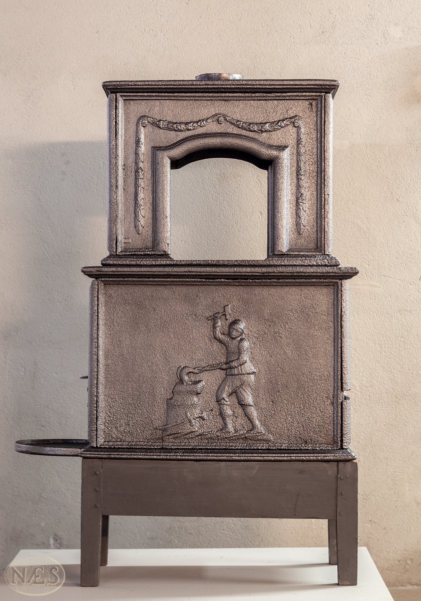 Etasjeovn med ovnsluke på kortsiden og røykuttak i toppen. På langsideplaten 1. etg en smed som holder en tang og hammer. En ambolt og et sverd liggende på bakken. Langside 2. etg har en buet åpning med girlande hengende rundt. Kortsiden 1. etg har en musling øverst. På kortsidene 2. etg et overflødighetshorn. Alle flatene er glate.