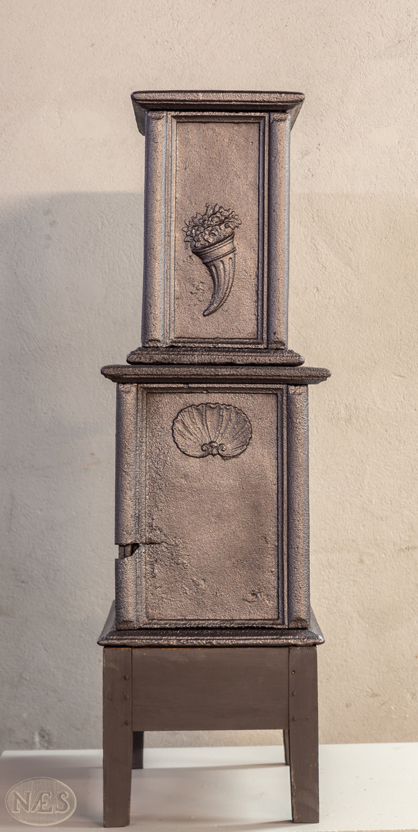 Etasjeovn med ovnsluke på kortsiden og røykuttak i toppen. På langsideplaten 1. etg en smed som holder en tang og hammer. En ambolt og et sverd liggende på bakken. Langside 2. etg har en buet åpning med girlande hengende rundt. Kortsiden 1. etg har en musling øverst. På kortsidene 2. etg et overflødighetshorn. Alle flatene er glate.