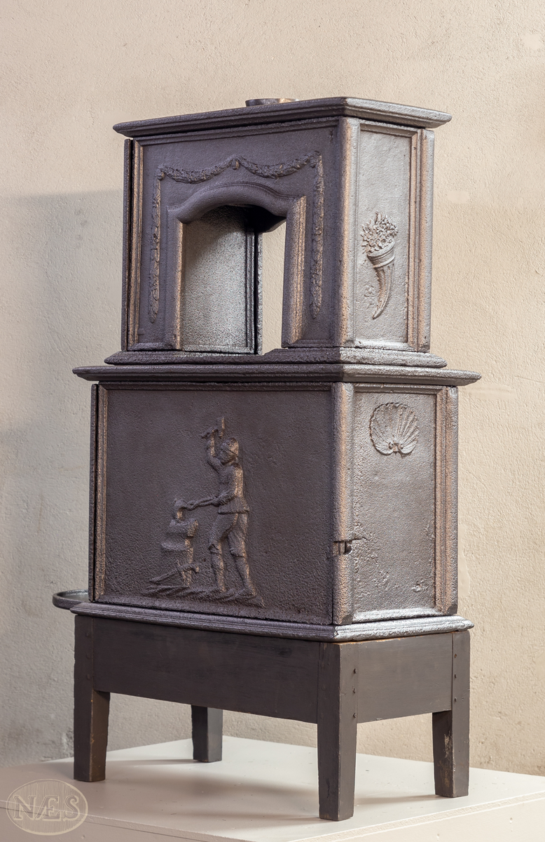 Etasjeovn med ovnsluke på kortsiden og røykuttak i toppen. På langsideplaten 1. etg en smed som holder en tang og hammer. En ambolt og et sverd liggende på bakken. Langside 2. etg har en buet åpning med girlande hengende rundt. Kortsiden 1. etg har en musling øverst. På kortsidene 2. etg et overflødighetshorn. Alle flatene er glate.