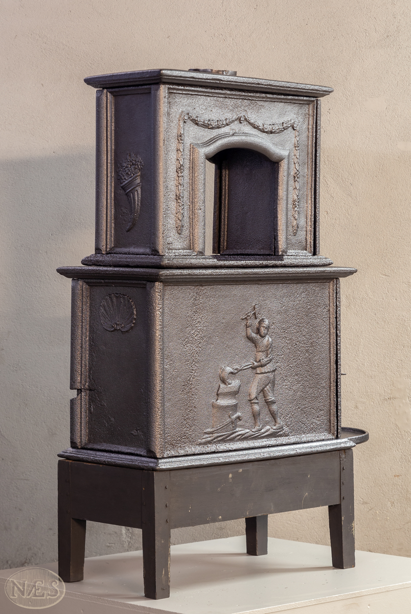 Etasjeovn med ovnsluke på kortsiden og røykuttak i toppen. På langsideplaten 1. etg en smed som holder en tang og hammer. En ambolt og et sverd liggende på bakken. Langside 2. etg har en buet åpning med girlande hengende rundt. Kortsiden 1. etg har en musling øverst. På kortsidene 2. etg et overflødighetshorn. Alle flatene er glate.