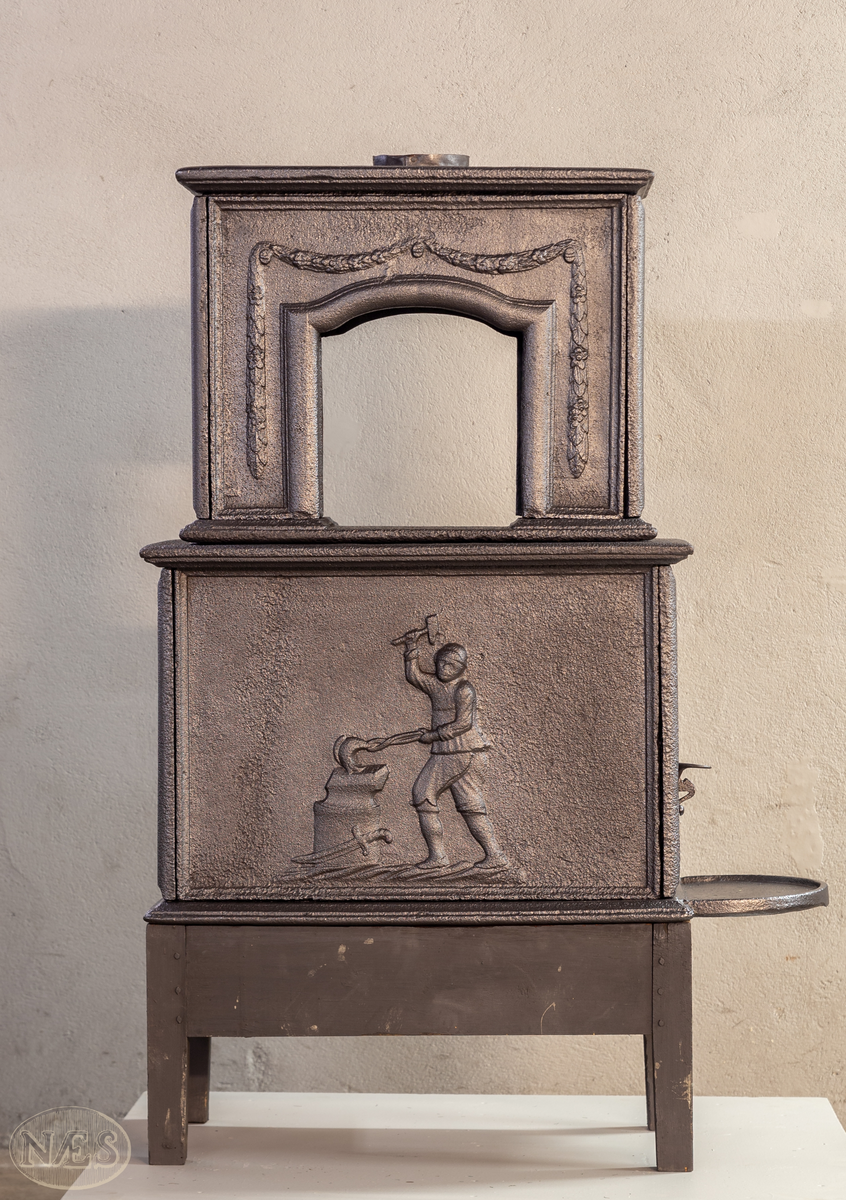 Etasjeovn med ovnsluke på kortsiden og røykuttak i toppen. På langsideplaten 1. etg en smed som holder en tang og hammer. En ambolt og et sverd liggende på bakken. Langside 2. etg har en buet åpning med girlande hengende rundt. Kortsiden 1. etg har en musling øverst. På kortsidene 2. etg et overflødighetshorn. Alle flatene er glate.