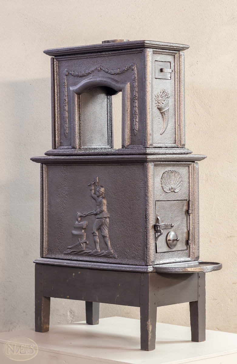 Etasjeovn med ovnsluke på kortsiden og røykuttak i toppen. På langsideplaten 1. etg en smed som holder en tang og hammer. En ambolt og et sverd liggende på bakken. Langside 2. etg har en buet åpning med girlande hengende rundt. Kortsiden 1. etg har en musling øverst. På kortsidene 2. etg et overflødighetshorn. Alle flatene er glate.