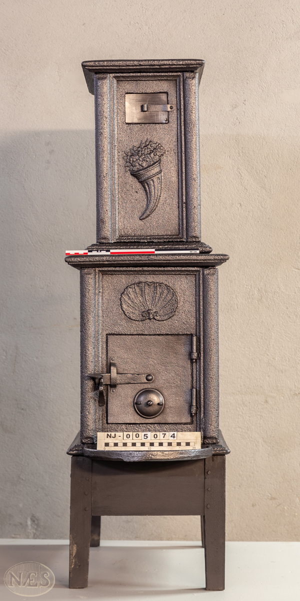 Etasjeovn med ovnsluke på kortsiden og røykuttak i toppen. På langsideplaten 1. etg en smed som holder en tang og hammer. En ambolt og et sverd liggende på bakken. Langside 2. etg har en buet åpning med girlande hengende rundt. Kortsiden 1. etg har en musling øverst. På kortsidene 2. etg et overflødighetshorn. Alle flatene er glate.