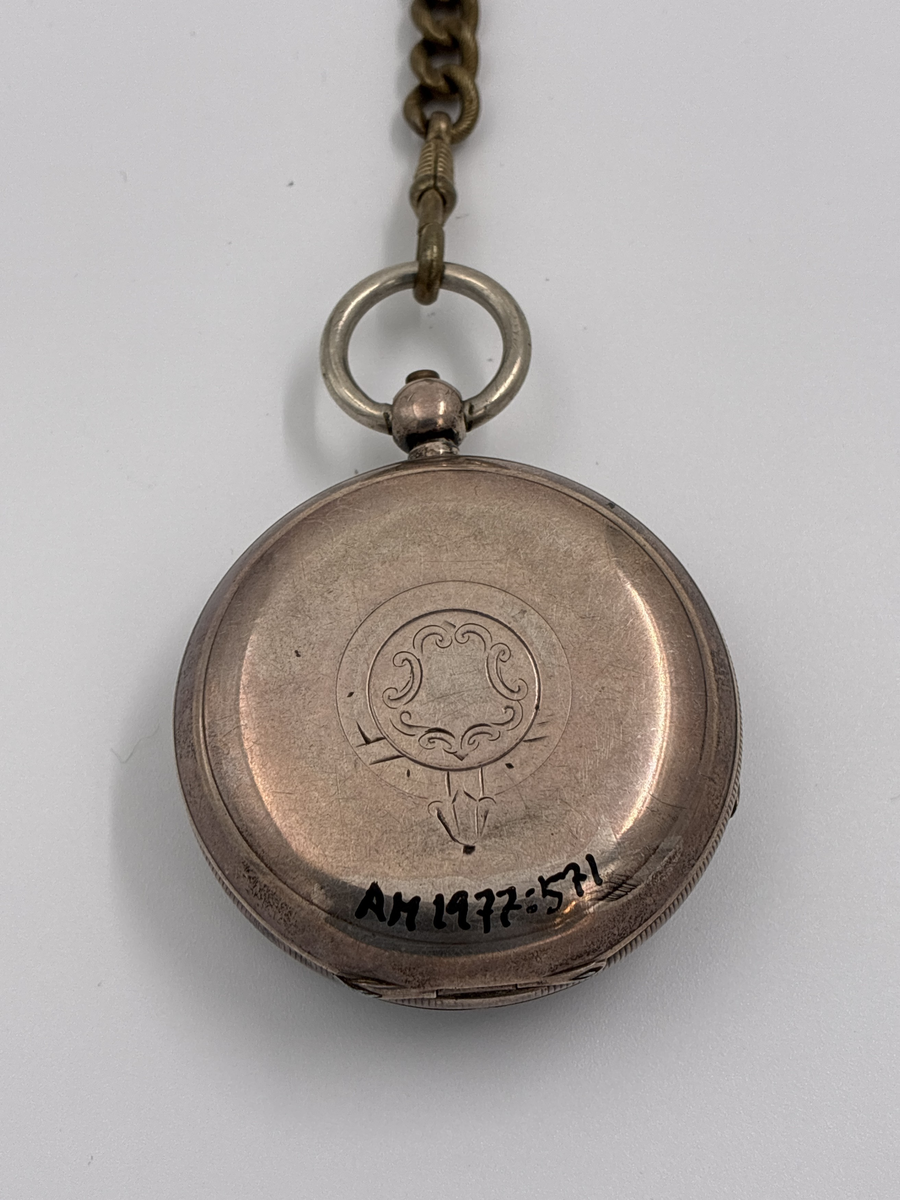 Fickur med vitemaljerad urtavla med romerska siffror och separat sekundvisare.
Text på urtavlan: "The Climax Trip Action Patent H. SAMUEL MANCHESTER".

Boett av silver med engelska stämplar
Lejon = England 
H.S = Okänd mästare 
Vapen med svärd och tre vetekärvar = Chester 
Otydbar årsstämpel

Kedja av mässing. Kedjans längd: ca 33 cm.