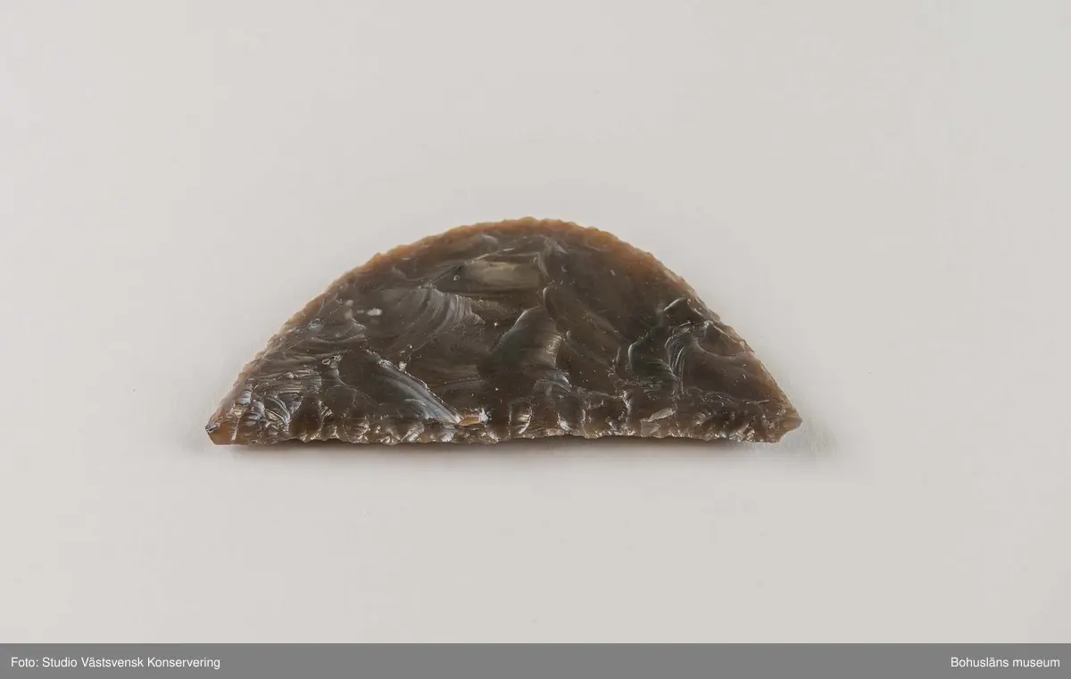 Skära, flathuggen, intakt, halvmåneformad. Brun något opak flinta. Användes vid skördearbete. Tydlig glans (gloss) längs eggdelen. Ett mycket fint och välbevarat exemplar. Denna typ hör till yngre stenålder - äldre bronsålder och kan vara avsiktligt nedlagd som depåfynd (Pedersen, 1993, s.139).