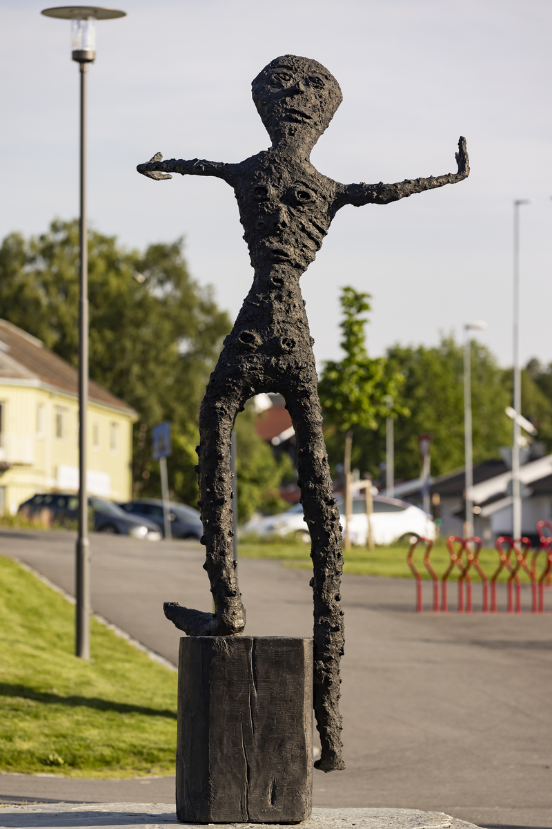 En gutt som balanserer på ett bein på en stubbe. En fot er kort, den andre er lang, armene er strekt forover. Figuren har mange ansikter, flere på baksiden. 