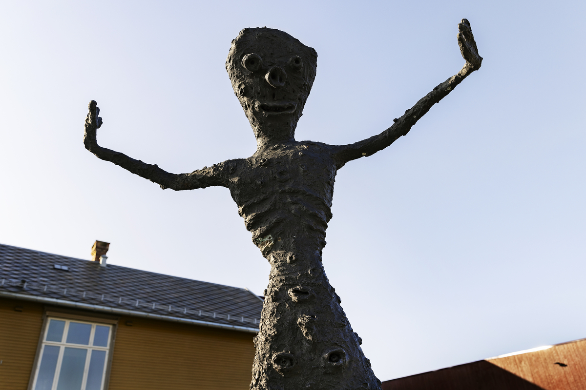 En gutt som balanserer på ett bein på en stubbe. En fot er kort, den andre er lang, armene er strekt forover. Figuren har mange ansikter, flere på baksiden. 