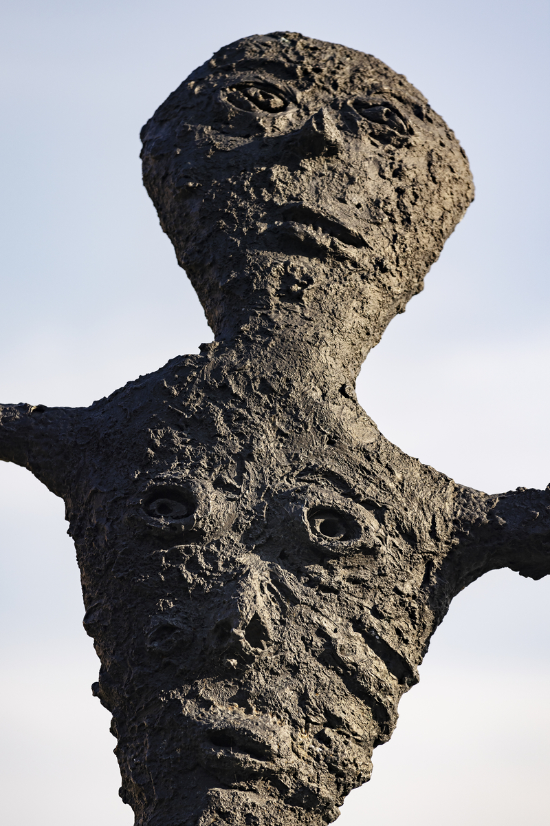 En gutt som balanserer på ett bein på en stubbe. En fot er kort, den andre er lang, armene er strekt forover. Figuren har mange ansikter, flere på baksiden. 