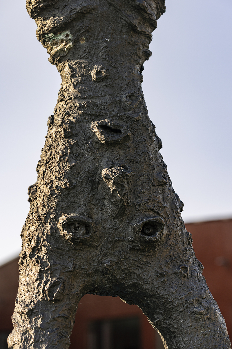 En gutt som balanserer på ett bein på en stubbe. En fot er kort, den andre er lang, armene er strekt forover. Figuren har mange ansikter, flere på baksiden. 