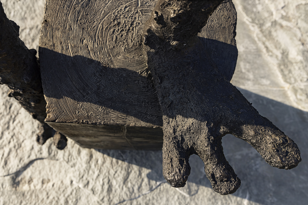 En gutt som balanserer på ett bein på en stubbe. En fot er kort, den andre er lang, armene er strekt forover. Figuren har mange ansikter, flere på baksiden. 