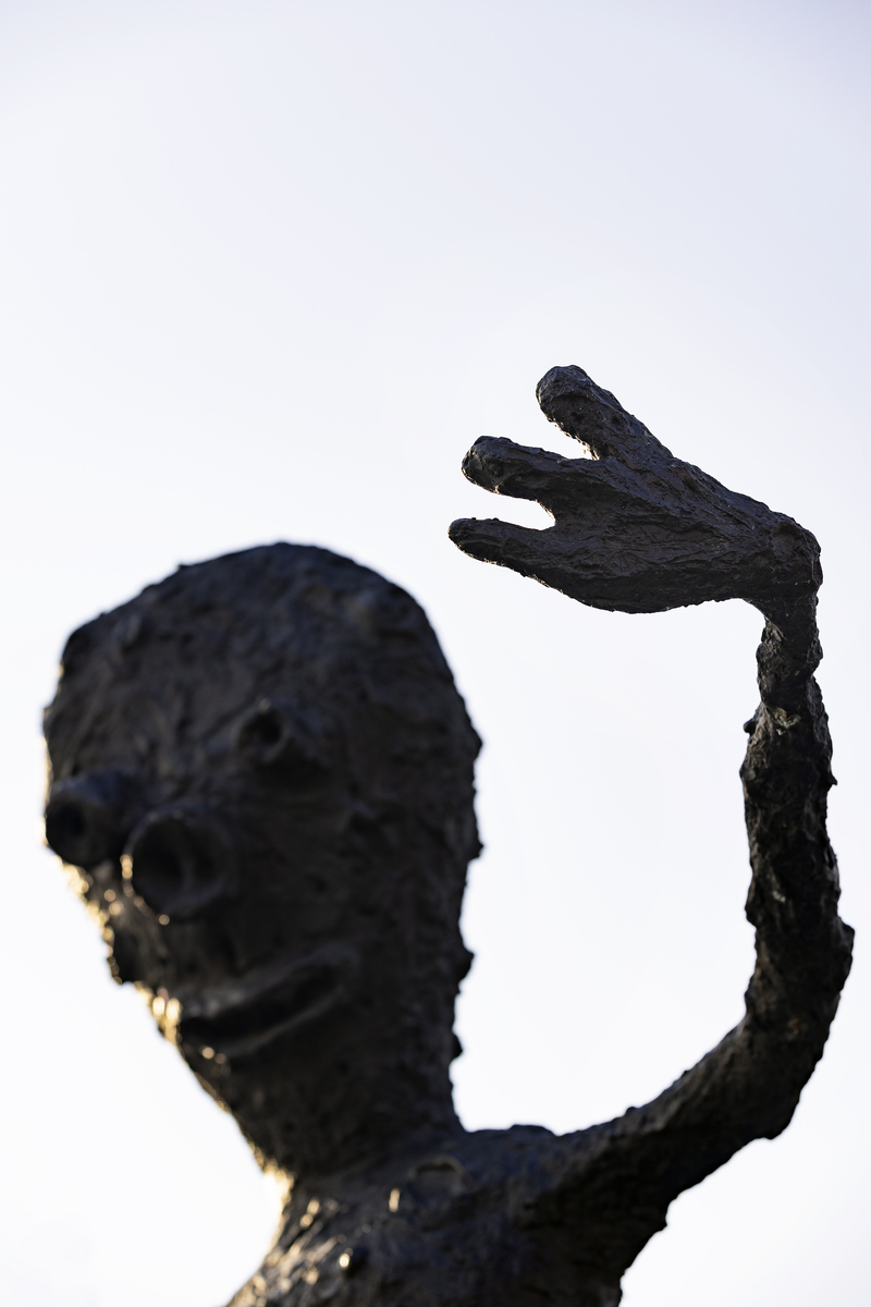 En gutt som balanserer på ett bein på en stubbe. En fot er kort, den andre er lang, armene er strekt forover. Figuren har mange ansikter, flere på baksiden. 