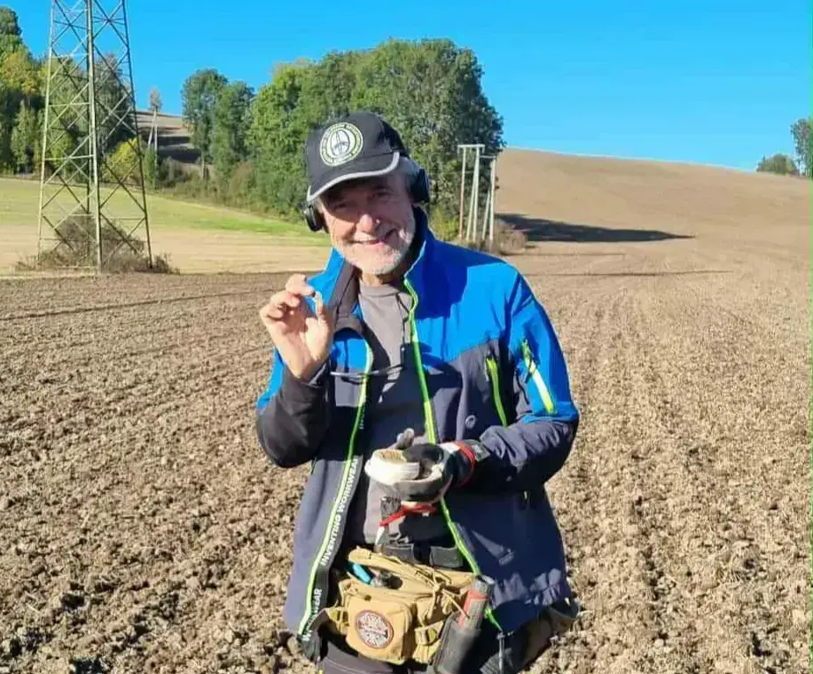 Metalldetektorist på et pløyd jorde viser fram et funn. 