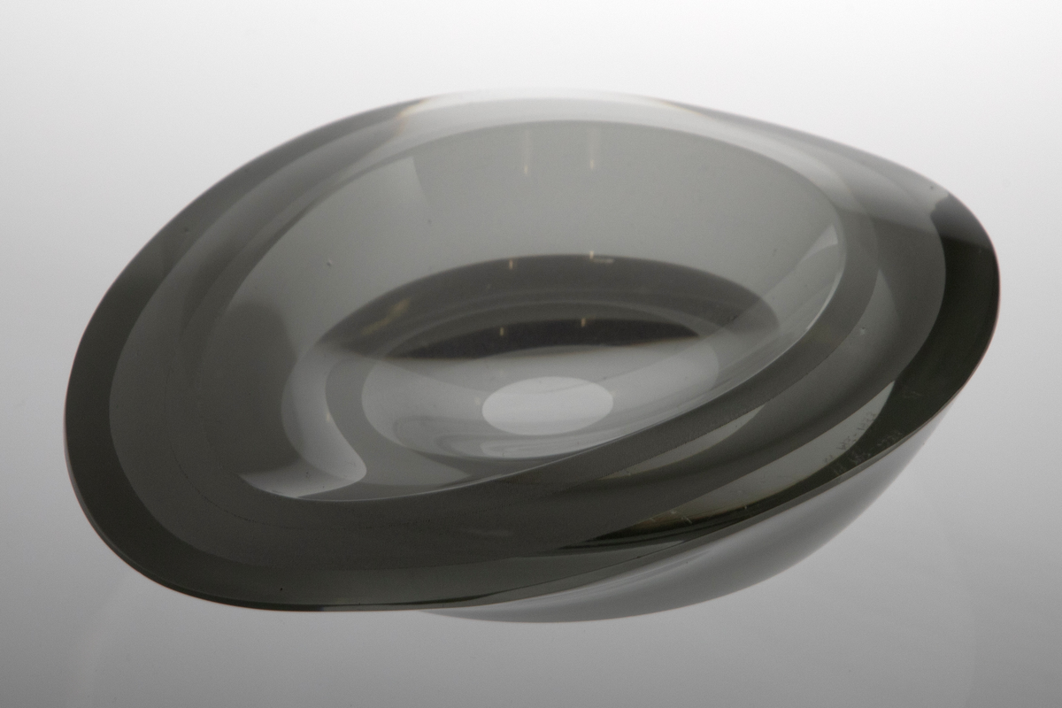 Askebeger i overfangsglass. Oval form med tilspisning i en side og en lav fordypning i midten. Sammensatt av tre tykke parallelle lag klart glass med matte mellomvegger. Lagene er skrådd inn mot bunnflaten og gir den midtre fordypningen sin form. Bunnflaten er dekorert med konsentriske ringer i variert matthetsgrad.