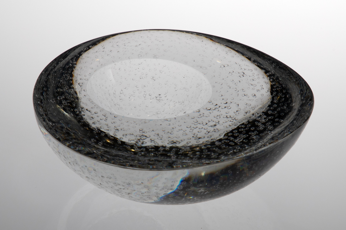 Askebeger i klart glass. Halvkuleformet med noe uregelmessig rund form øverst. Avrundet bunnflate. På overflaten en mindre halvkuleformet fordypning, plassert noe bort fra flatens sentrum. Utsiden er etterbehandlet med punsling.