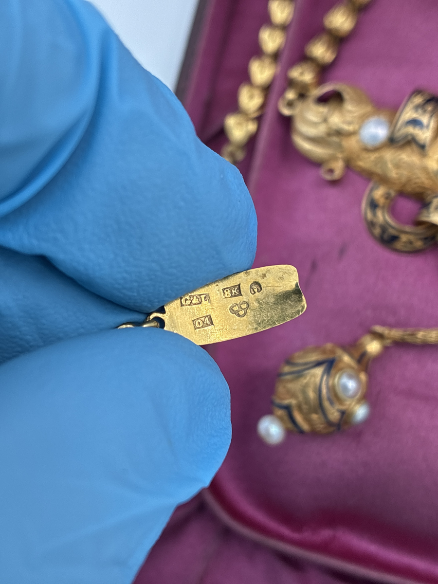 Halssmycke av guld. Smycket med graverad dekor, delvis med mörkblå emalj. Fyra infattade pärlor. Två runda kläppar med vardera fyra små pärlor. I etui, ej ursprungligen sammanhörande. 

Stämplar:
Trekronorsstämpel
Årsbokstav: O4 =1844
Guldhalt = 18K
Stadsstämpel: S:t Erik =Stockholm
Mästarstämpel: G&L = Giron & Löngren: Marc Giron och Olof Wilhelm Löngren, Stämplade gemensamt 1820-1847. Giron var svärfar till Löngren. 

Storlek: Längd med kedja 56,2 cm.