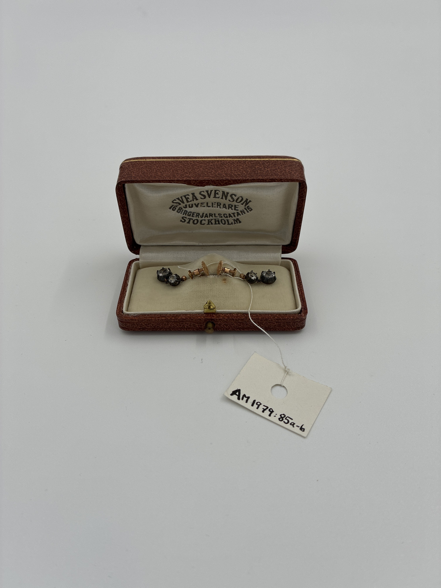 Ett par örhängen av guld med två hängande vita stenar, infattade i silver, i vardera.
 
Stämplar: 
Trekronorsstämpel
Årsbokstav: H7= 1910
Stadsstämpel: S:t Erik (=Stockholm)
"18 K"
Mästarstämpel: "JP"