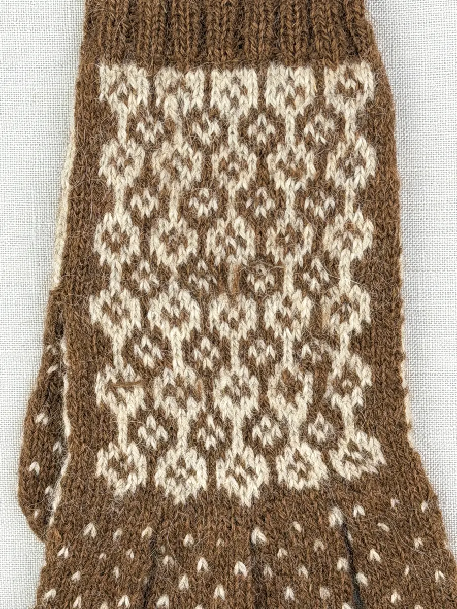 En stickad fingervante, med randig mudd i 2-2 resår (2 räta och 2 aviga maskor) och mönsterstickad vante i slätstickning, av 2-trådigt ullgarn.