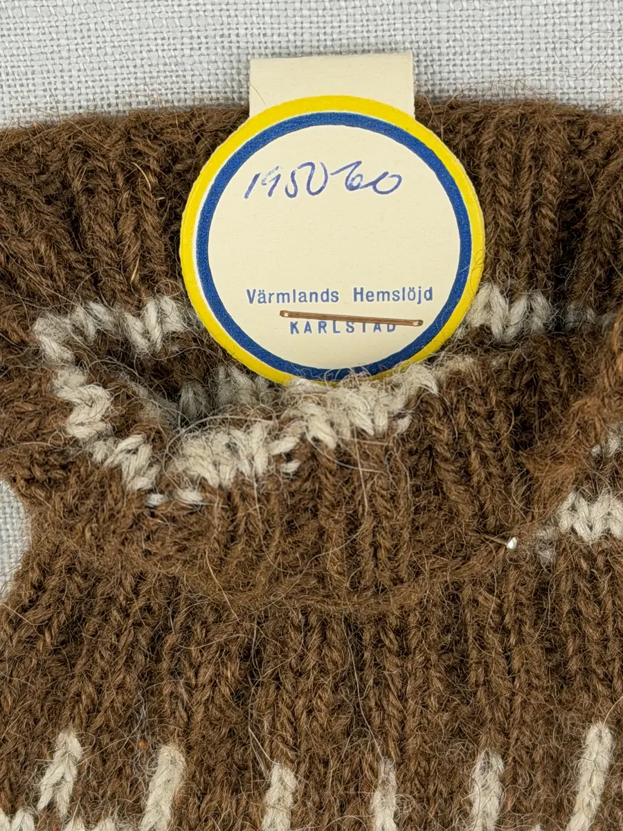 En stickad fingervante, med randig mudd i 2-2 resår (2 räta och 2 aviga maskor) och mönsterstickad vante i slätstickning, av 2-trådigt ullgarn.