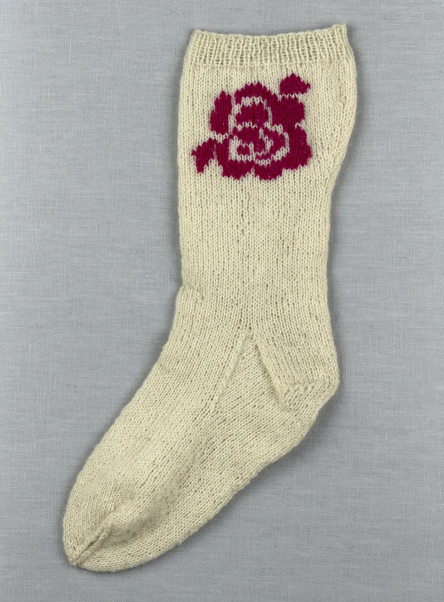 En stickad socka, med mudd i 1-1 resår (1 rät och 1 avig maska) och skaft och fot i slätstickning av 2-trådigt ullgarn. Sockans skaft är dekorerad på yttersidan med ett ullgarns broderi i maskstygn.
