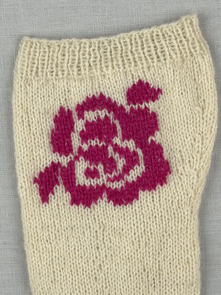 En stickad socka, med mudd i 1-1 resår (1 rät och 1 avig maska) och skaft och fot i slätstickning av 2-trådigt ullgarn. Sockans skaft är dekorerad på yttersidan med ett ullgarns broderi i maskstygn.
