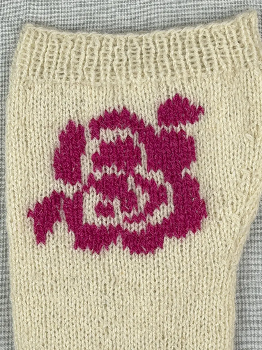 En stickad socka, med mudd i 1-1 resår (1 rät och 1 avig maska) och skaft och fot i slätstickning av 2-trådigt ullgarn. Sockans skaft är dekorerad på yttersidan med ett ullgarns broderi i maskstygn.
