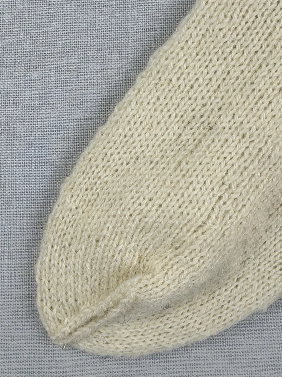 En stickad socka, med mudd i 1-1 resår (1 rät och 1 avig maska) och skaft och fot i slätstickning av 2-trådigt ullgarn. Sockans skaft är dekorerad på yttersidan med ett ullgarns broderi i maskstygn.
