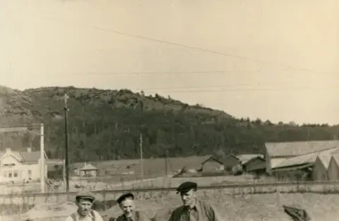Fotografi taget av Torsten Nilsson som var anställd hos Grens handelsträdgård c:a 1945 - 1960 och expert på nejlikor. 

Tre män som bär keps står i förgrunden, medan berg och skog skymtar i bakgrunden, tillsammans med en övergång vid järnvägen. 

Osäkert när fotografiet kan vara taget.