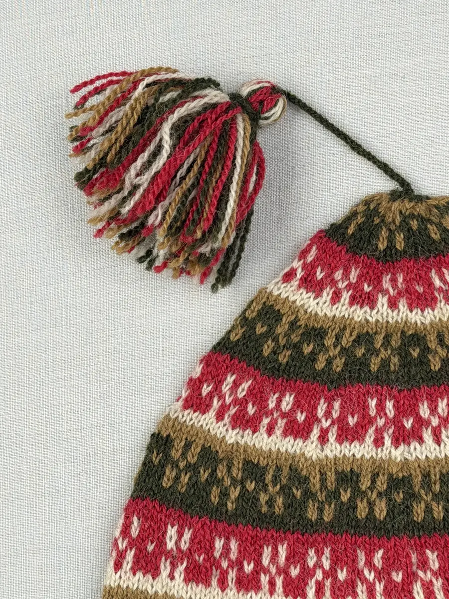 En stickad mössa, i mönsterstickad slätstickning, av 2-trådigt ullgarn. Mössan är rundstickad och har en fåll med 1-1 resår (1 rät och 1 avig maska) i nederkant, som är fastsydd med kaststygn. Toppen på mössan är formad med strålhoptagning och högst upp sitter en tofs fastsatt med en påtad snodd.
