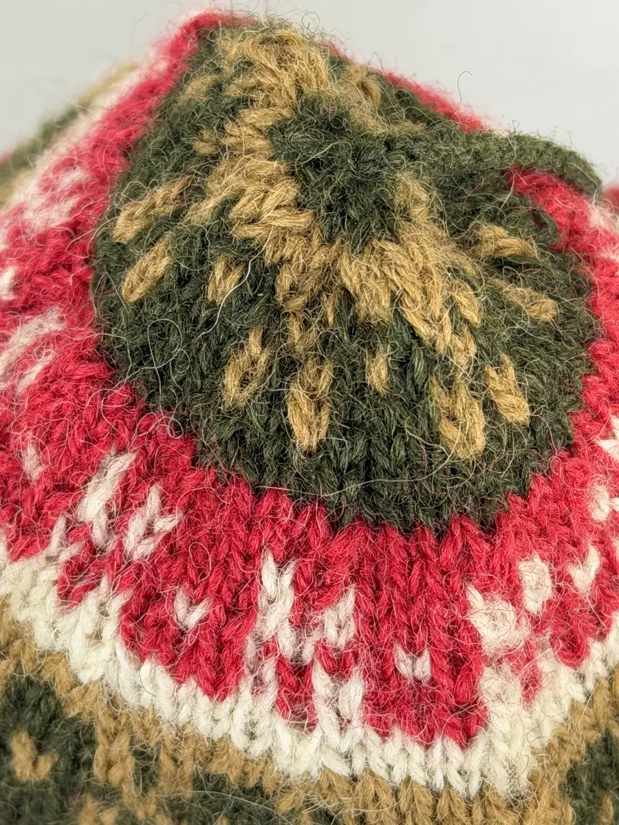 En stickad mössa, i mönsterstickad slätstickning, av 2-trådigt ullgarn. Mössan är rundstickad och har en fåll med 1-1 resår (1 rät och 1 avig maska) i nederkant, som är fastsydd med kaststygn. Toppen på mössan är formad med strålhoptagning och högst upp sitter en tofs fastsatt med en påtad snodd.
