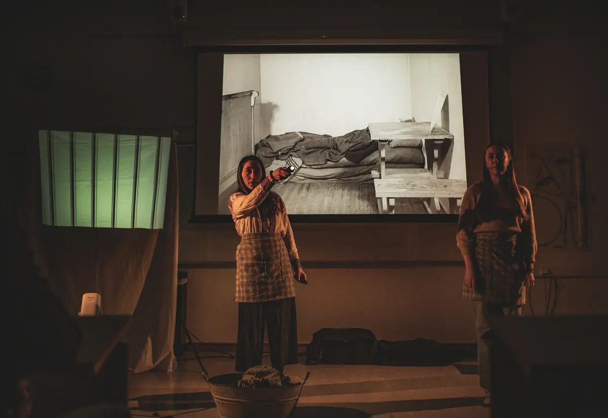 Ei jente står med forkle og peker ei lommelykt ut mot et publikum, ved siden av henne står ei anna jente og følger med