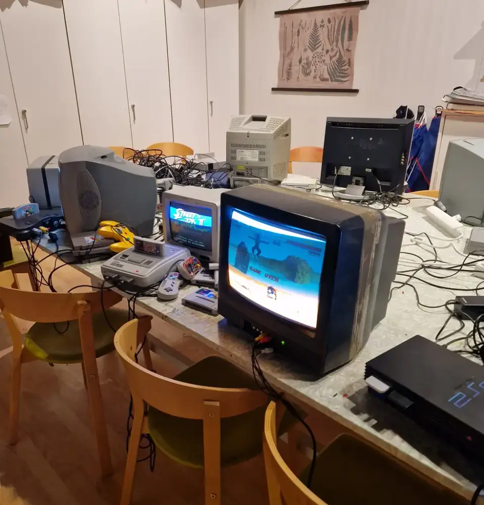 Flera gammeldags datorer med spelkonsoler och handkontroller av olika slag står uppställda på ett bord med skåp i bakgrunden.