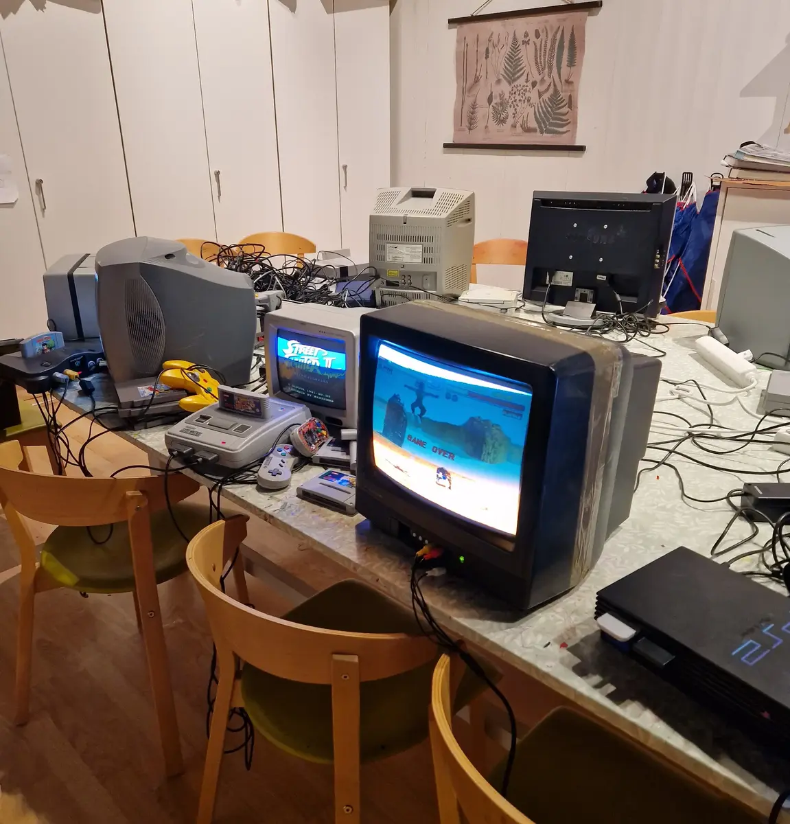 Flera gammeldags datorer med spelkonsoler och handkontroller av olika slag står uppställda på ett bord med skåp i bakgrunden.