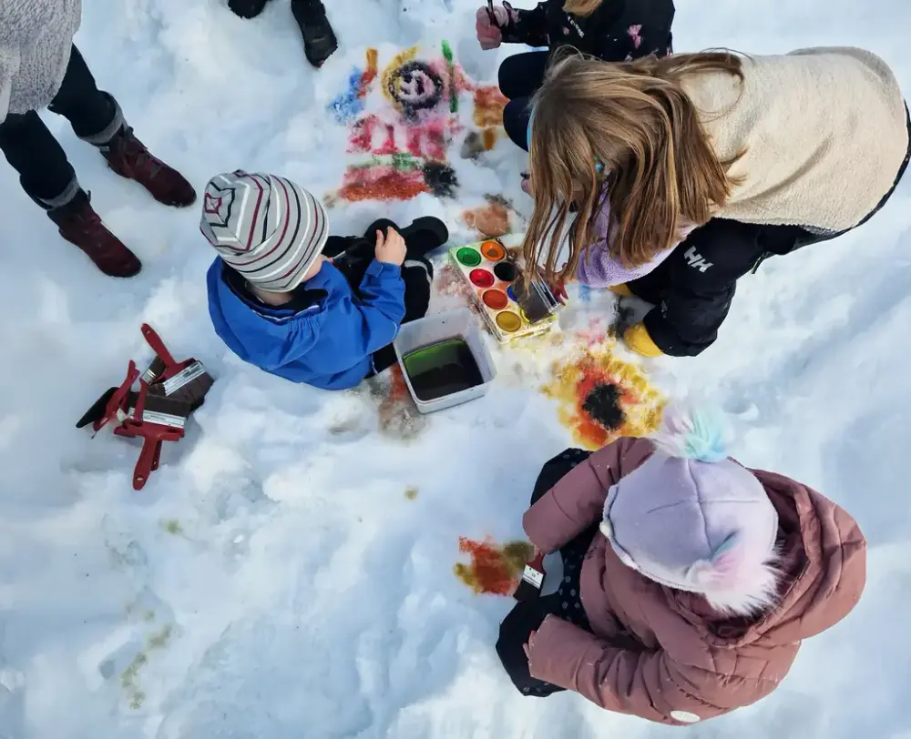 Barn med jackor och mössor sitter i snön och målar olika geometriska figurer i olika färger.