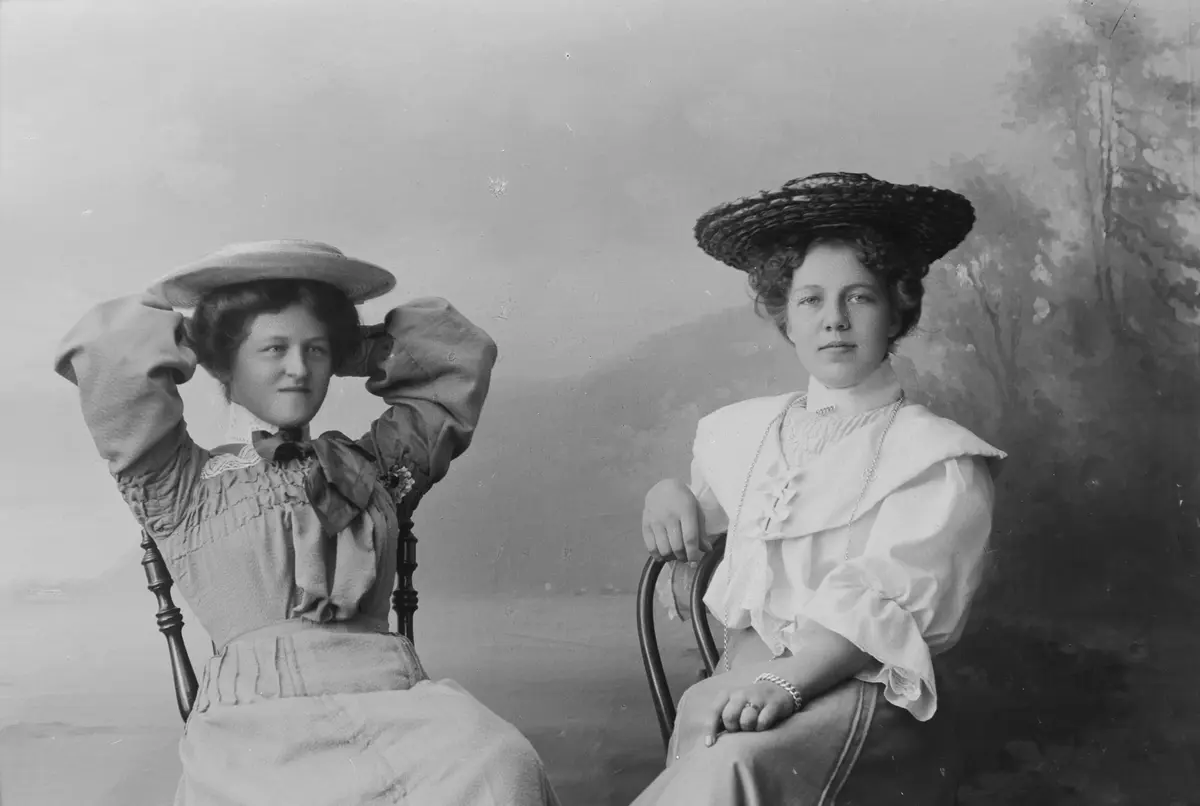 Två kvinnor i hattar och klänningar sitter på varsin stol.