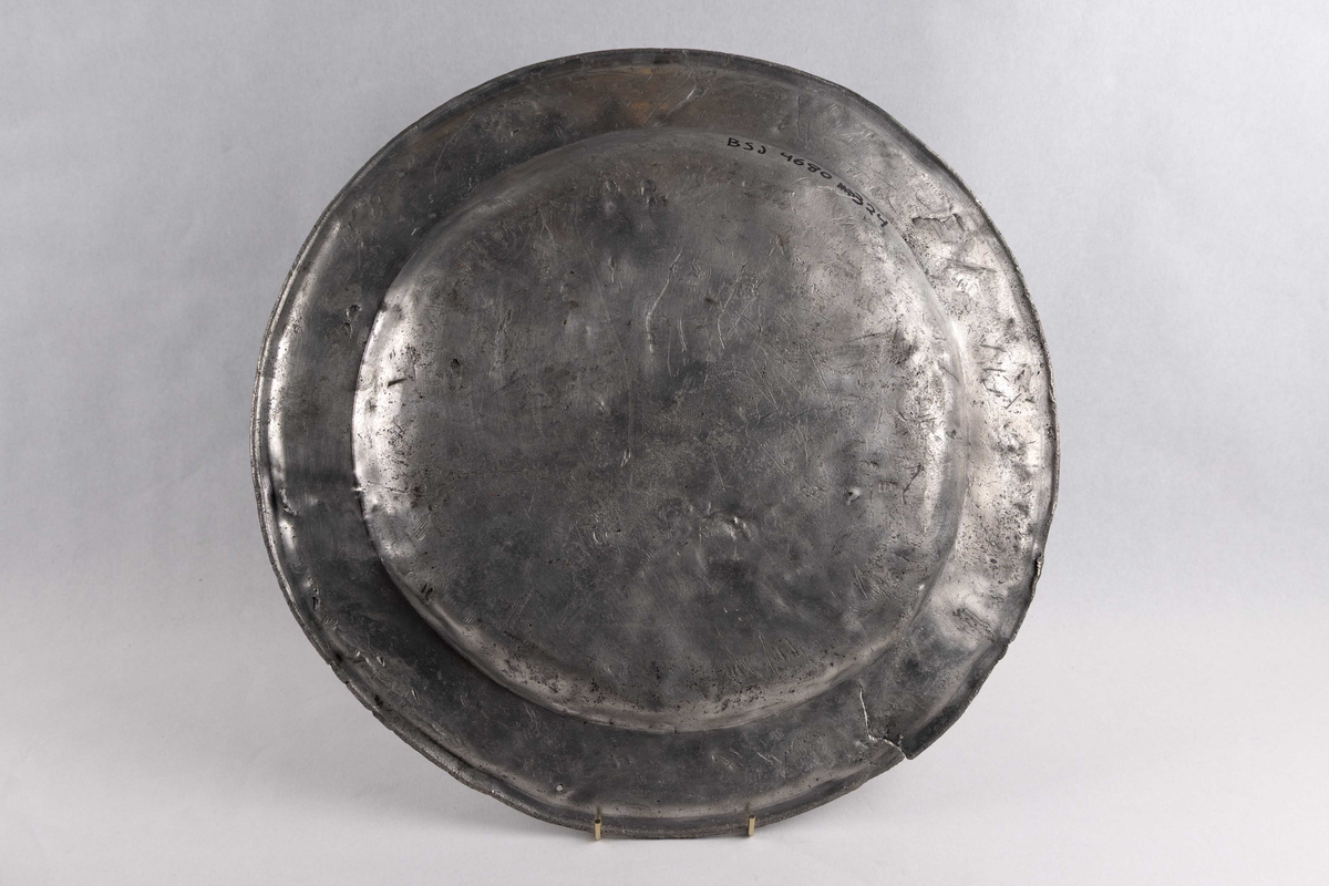 Stort, sirkulært serveringsbrett funnet ved skipet AKERENDAM som forliste på Runde i 1725. Tinnfat med flat bunn og 5 cm. bred rand. To stempler på undersiden. Motiv er en engel med vinger, holder noe i hendene. Det andre stempelet er blomst og krone. Brett med bulker/skader.