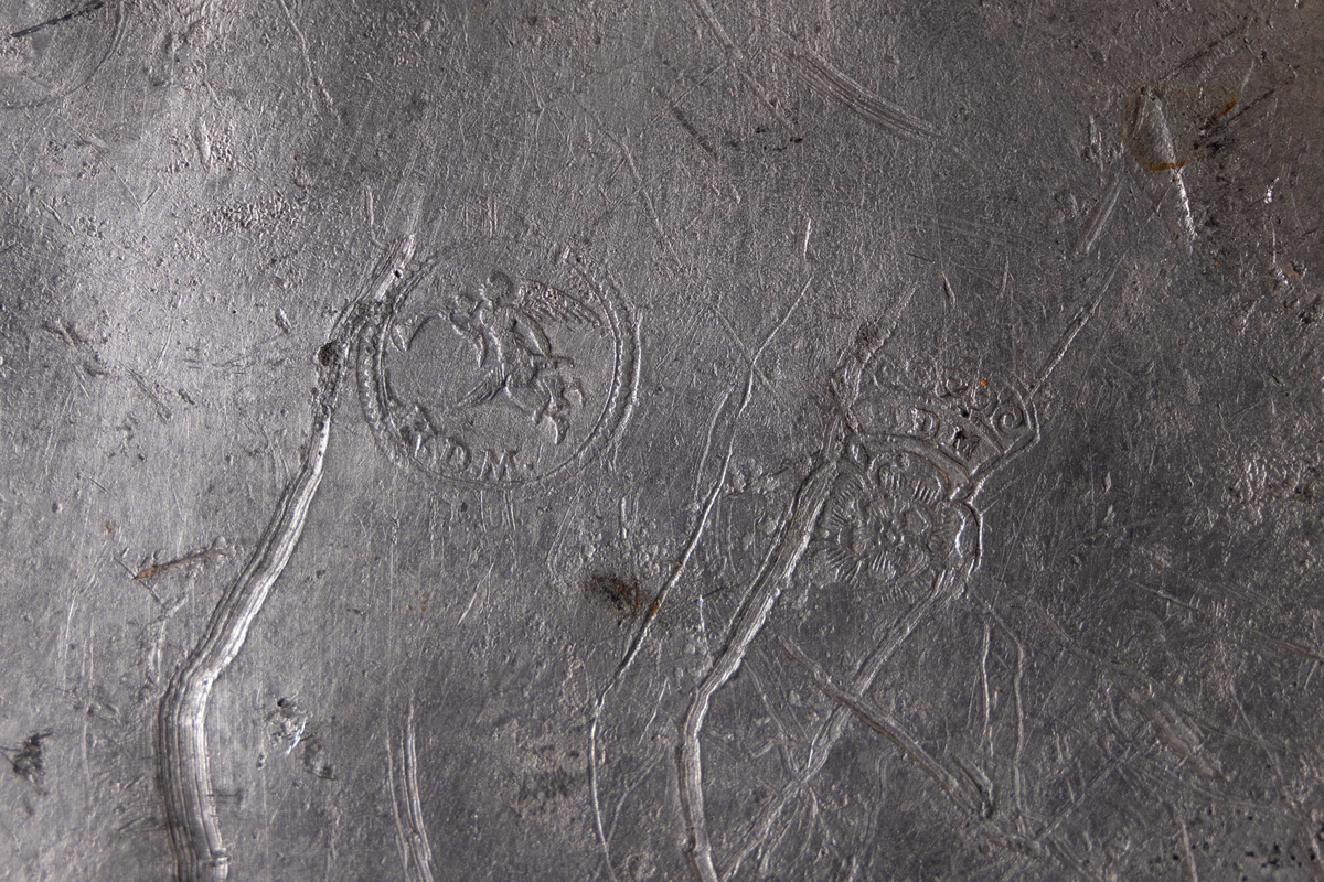 Stort, sirkulært serveringsbrett funnet ved skipet AKERENDAM som forliste på Runde i 1725. Tinnfat med flat bunn og 5 cm. bred rand. To stempler på undersiden. Motiv er en engel med vinger, holder noe i hendene. Det andre stempelet er blomst og krone. Brett med bulker/skader.