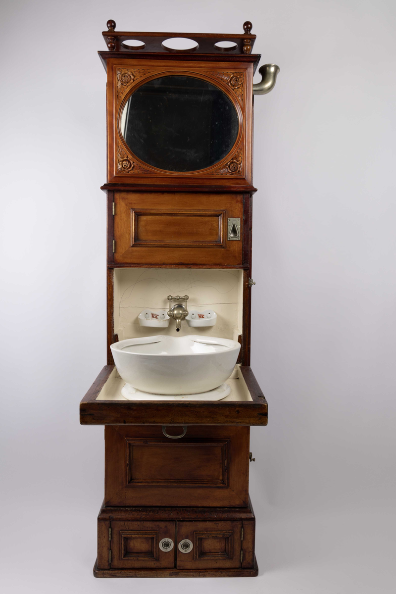 Servantskap fra BDS skip. Skap med vannbeholder og oppsamlingstankt samt servant som kan felles ned ved bruk. Speil på øvre skapdør med treutskjæringer rundt som dekor. Bak speilet er det en beholder for vann. Under denne er en dør som skjuler ett skap inndelt i fire små rom. Så kommer 1 stk. dør som kan felles ned hvor det er montert en porselensvask. På veggen bak er det en vannkran samt to stk. sepeholdere med BDS logo. Under denne 1 stk. dør som skjuler oppsamlingstank i metall for vaskevann. Nederst mot gulv to stk. små dører som skjuler to mindre rom.