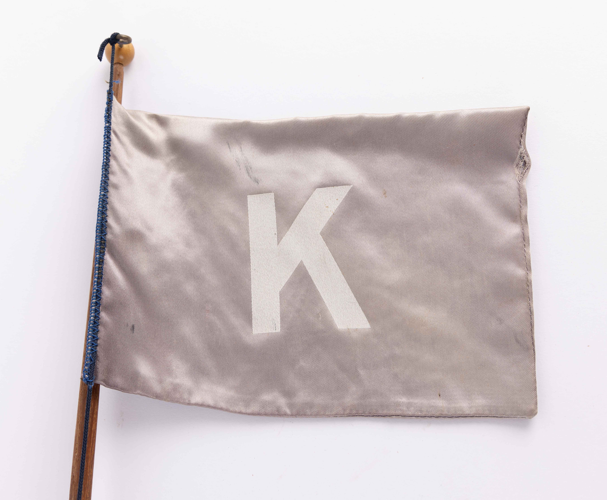 Bordvimpel fra Jacob Kjødes rederi i Bergen montert på flaggstang av brunt tre. Flagg med blå bunn og hvit bokstav (flagget er falmet).