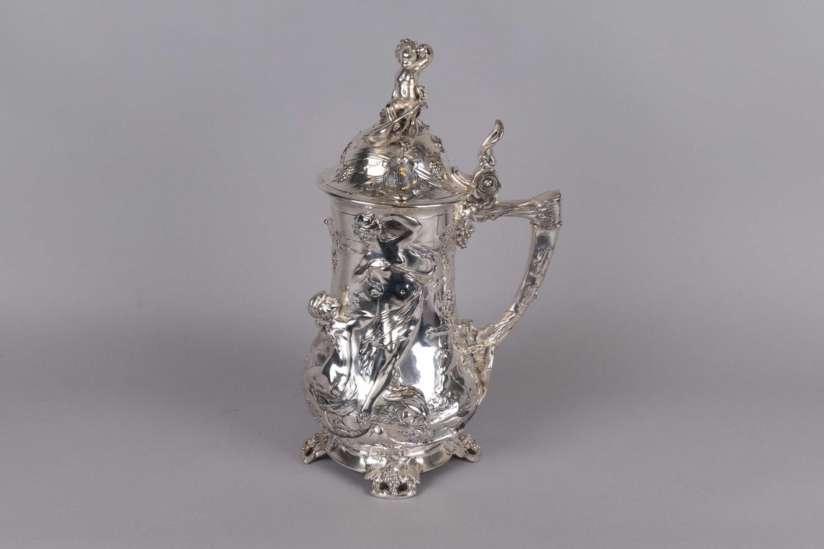 Kanne dekorert med gjennomgådene relieffmønster av figurer og kjeruber blant vinranker og druer. Kuppellokk med motiv av en kjerub med banjo som skåler med et glass vin. Håndtaket dekorert med vinranker og druer.