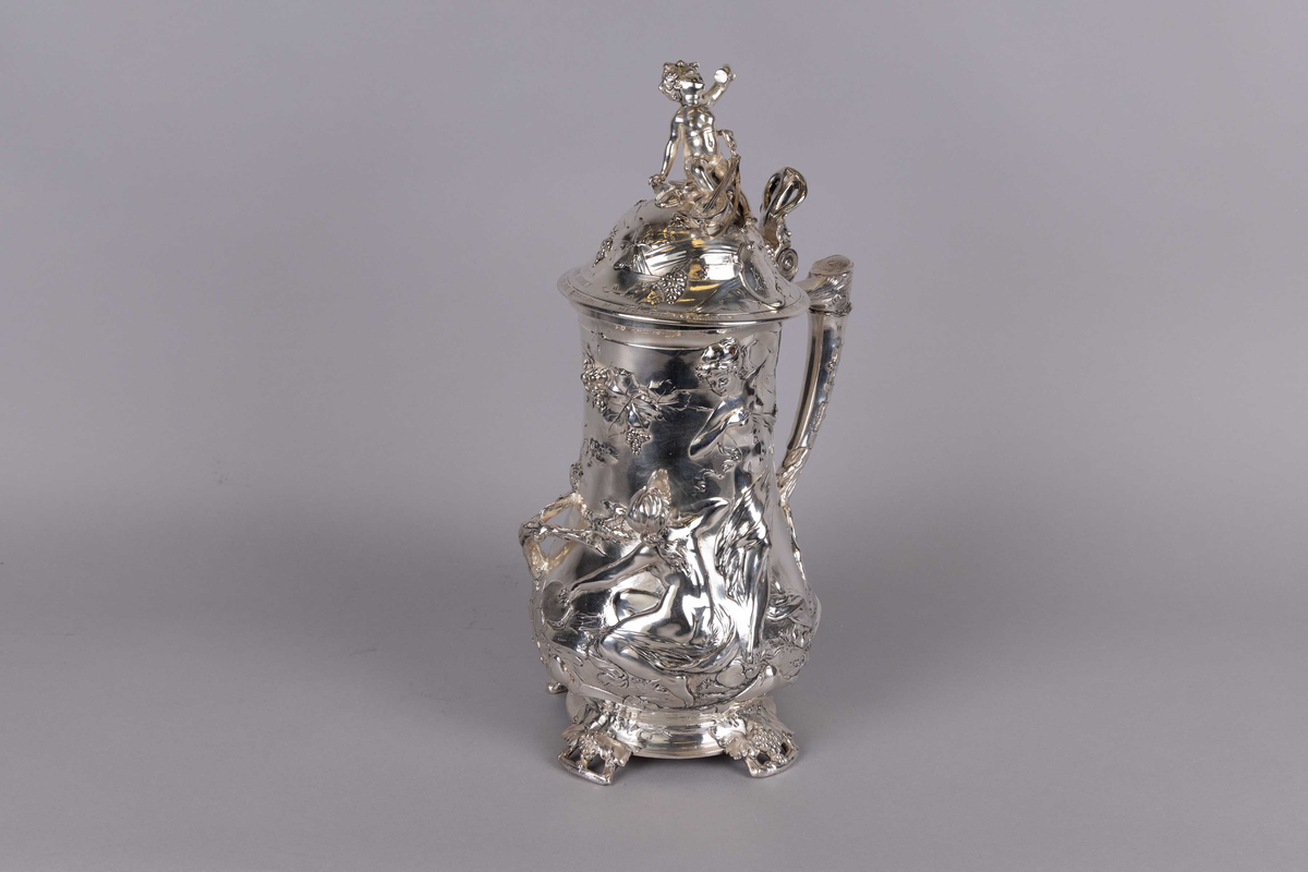 Kanne dekorert med gjennomgådene relieffmønster av figurer og kjeruber blant vinranker og druer. Kuppellokk med motiv av en kjerub med banjo som skåler med et glass vin. Håndtaket dekorert med vinranker og druer.