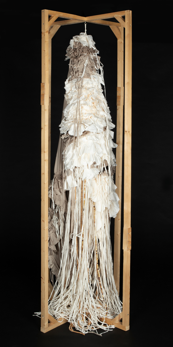 Tekstil skulptur i lyse farger som henger fra en frittstående firesidet ramme lagd av stenderverk i tre.