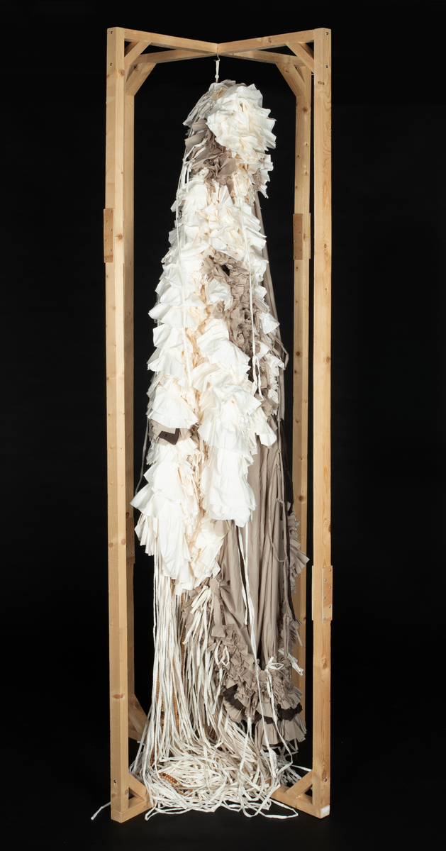 Tekstil skulptur i lyse farger som henger fra en frittstående firesidet ramme lagd av stenderverk i tre.