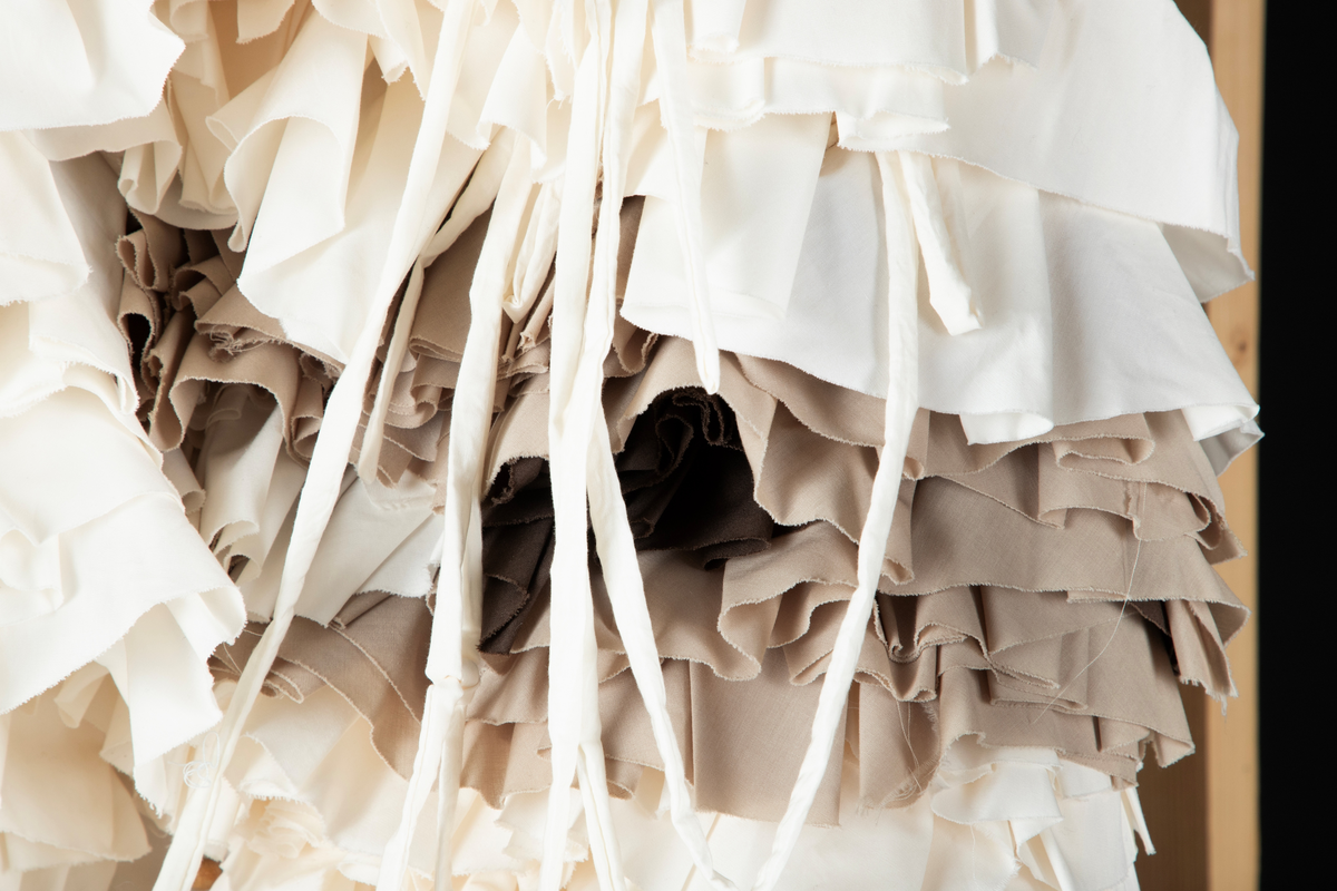 Tekstil skulptur i lyse farger som henger fra en frittstående firesidet ramme lagd av stenderverk i tre.