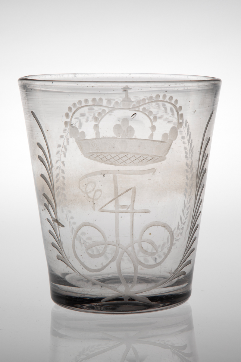 Beger i klart glass. Tilnærmet sylinderformet korpus med svakt koniske vegger i tykt glass. Yttersiden er dekorert med sliping og gravering. På den ene siden Fredrik IVs bekronede monogram, omgitt av to kryssende bladverk. På den andre siden en oval i form av en gravert stilisert plantebord som omkranser seks slipte sirkler. Rester etter puntemerke, noe grålig glassmasse.