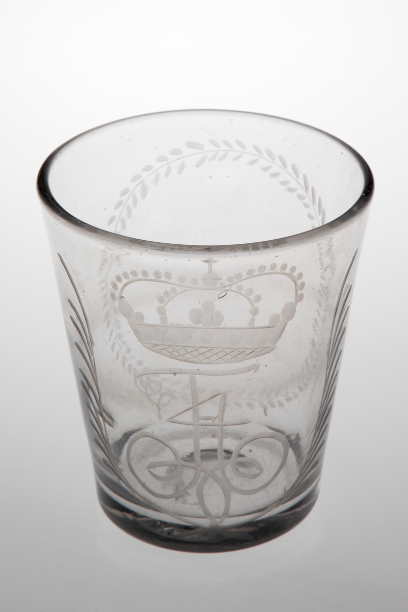 Beger i klart glass. Tilnærmet sylinderformet korpus med svakt koniske vegger i tykt glass. Yttersiden er dekorert med sliping og gravering. På den ene siden Fredrik IVs bekronede monogram, omgitt av to kryssende bladverk. På den andre siden en oval i form av en gravert stilisert plantebord som omkranser seks slipte sirkler. Rester etter puntemerke, noe grålig glassmasse.